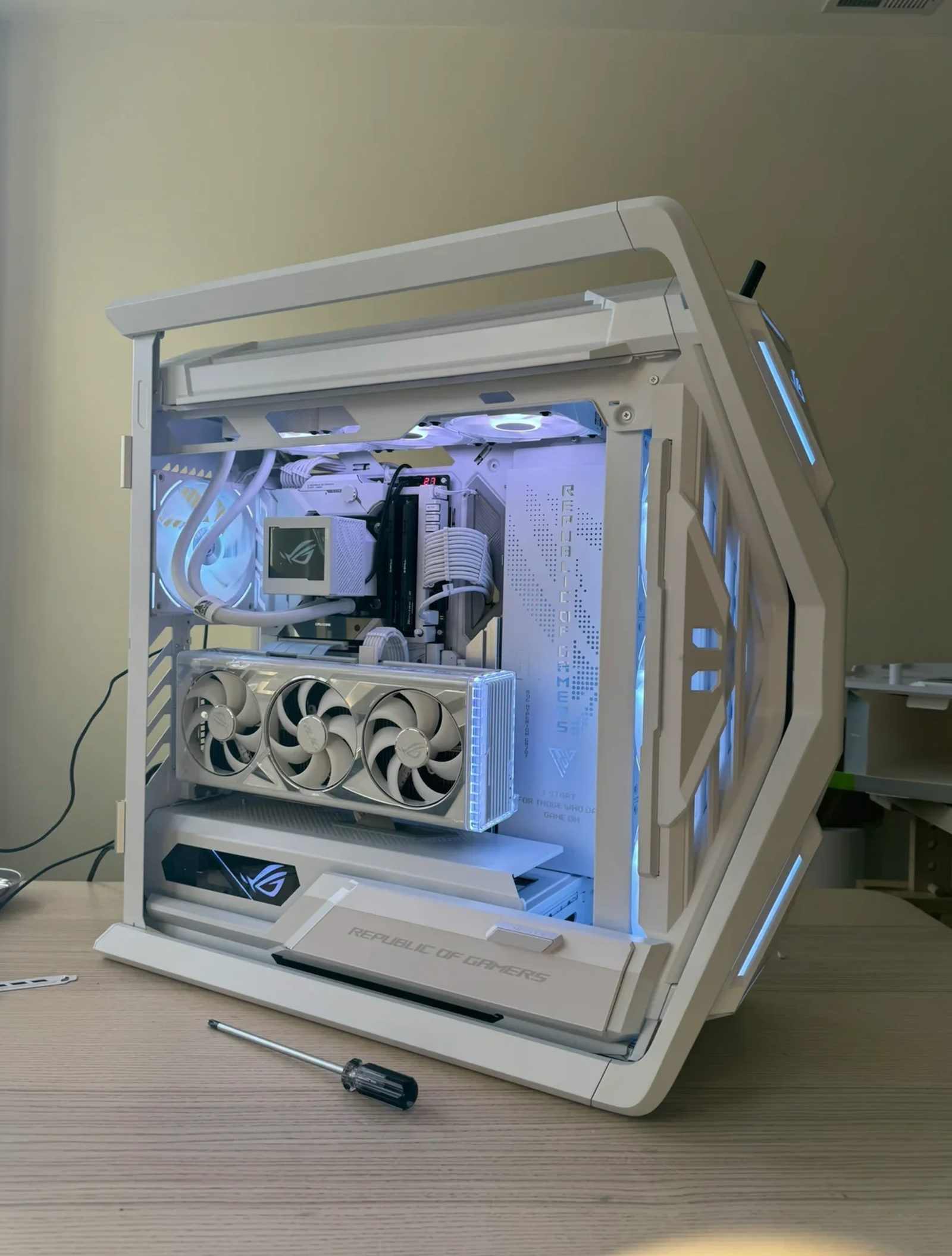 A white ASUS ROG case mid-build with its panels off, showing white cabling, a white AIO cooler and a white graphics card, a screwdriver lying on the desk beside it