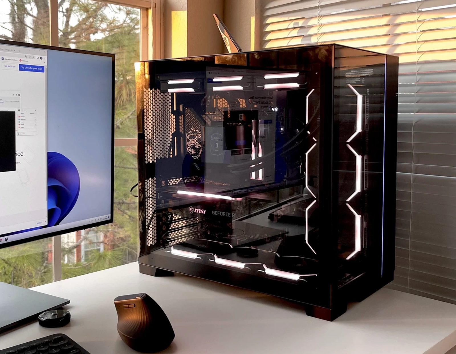 A black glass-paneled tower on a desk in daylight, lit strips glowing softly, next to a monitor and mouse