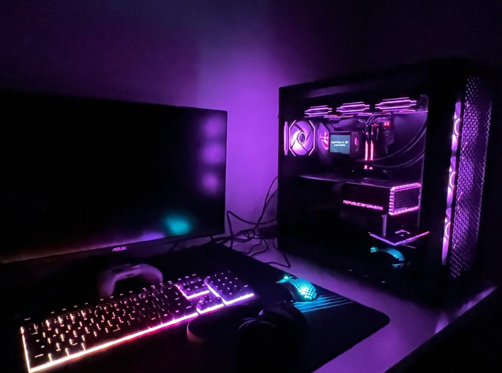A desk at night lit almost entirely by the machine: violet light across the wall, a glowing keyboard, and the case beside a dark monitor