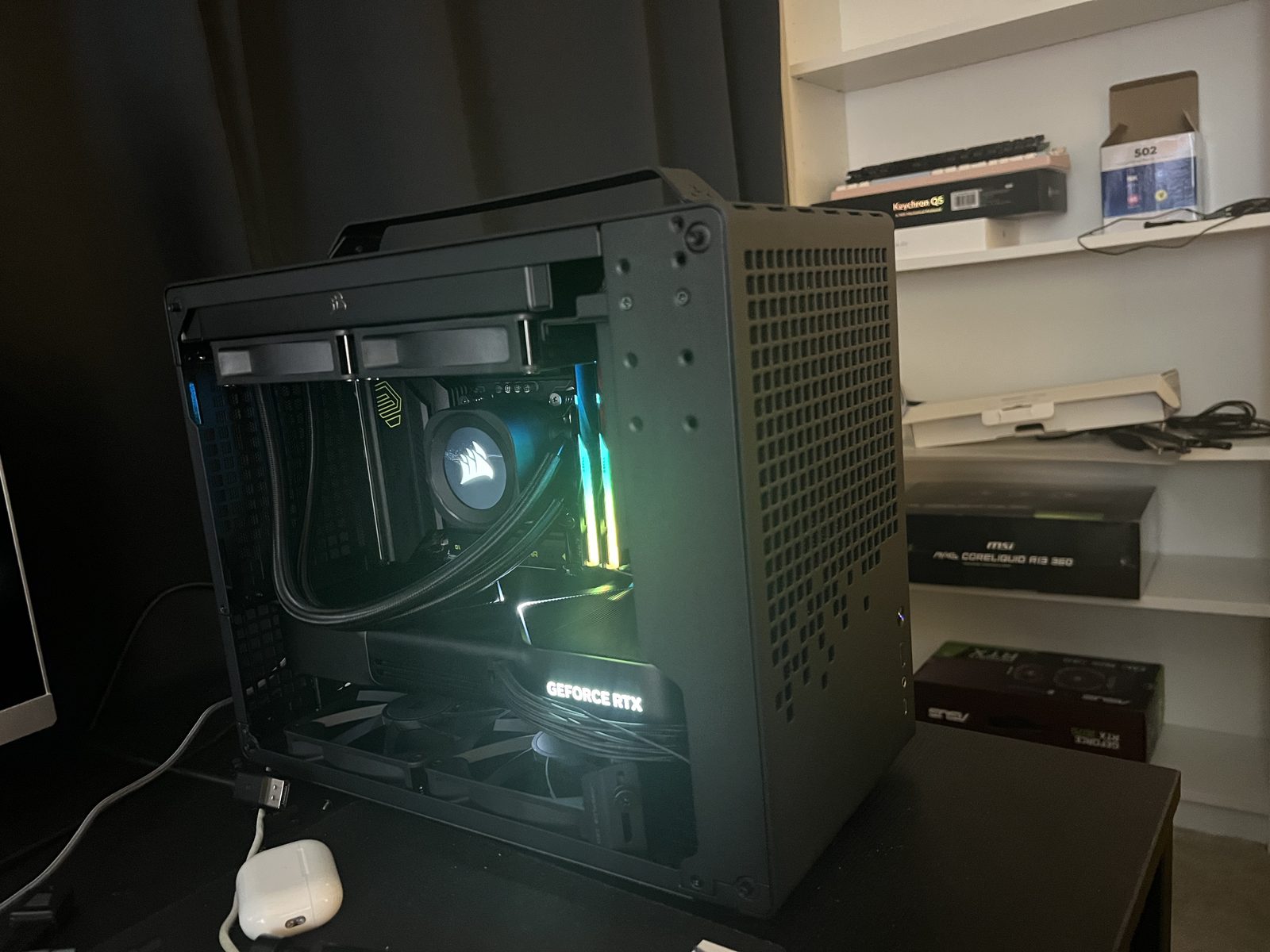 A small-form-factor black case on a desk, spare components and boxes on shelves behind it